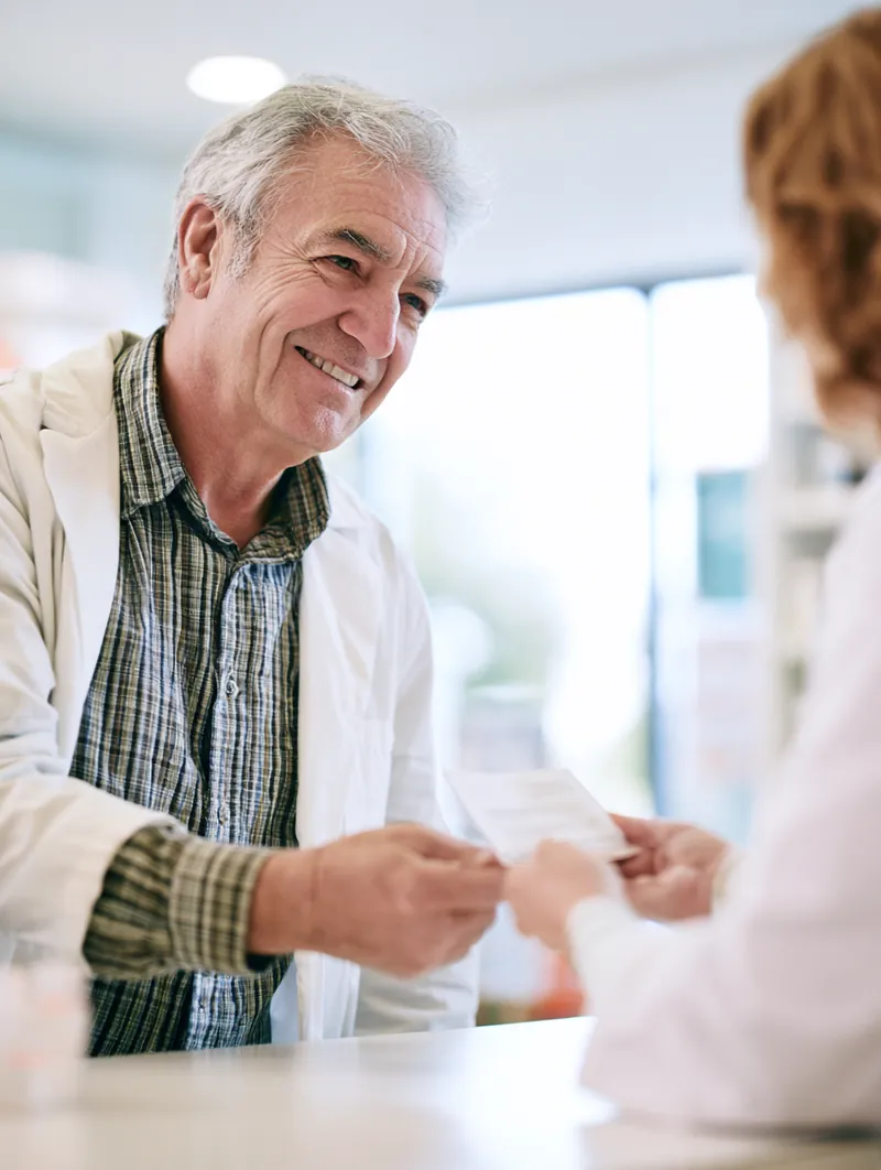 A pharmacist hands a small pharmacy package across the counter to a man in his 60s